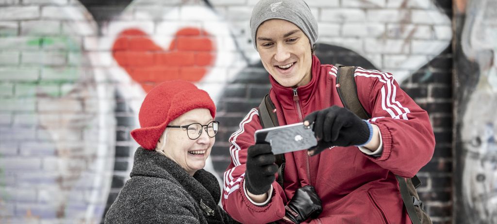 Vanha nainen ja nuori mies katsovat yhdessä kännykän ruutua ulkovaatteissa. Taustalla näkyy harmaalle seinälle maalattu punavalkoinen graffitisydän.