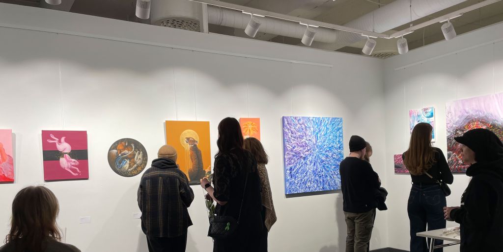Ihmisiä gallerian avajaisissa, katselemassa seinälle ripustettuja taideteoksia. Teokset ovat punaisen, sinisen ja oranssin sävyissä.