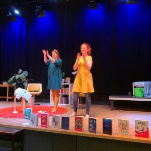 Kaksi naista teatterilavalla taputtamassa käsiään kohti yleisöä. Lavan etualalla useita kirjoja seisomassa kannet kohti yleisöä.