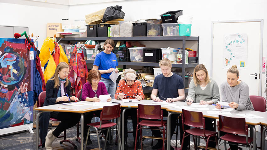 Kuusi henkilöä pöydän ääressä piirtämässä ja kirjoittamassa. Taustalla toiminnan ohjaaja. Ympäristönä on taidepaja.