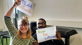 Kaksi henkilöä paperikylttien kanssa, joissa lukee 'Tilaa kulttuurille' ja Room for Cultures'