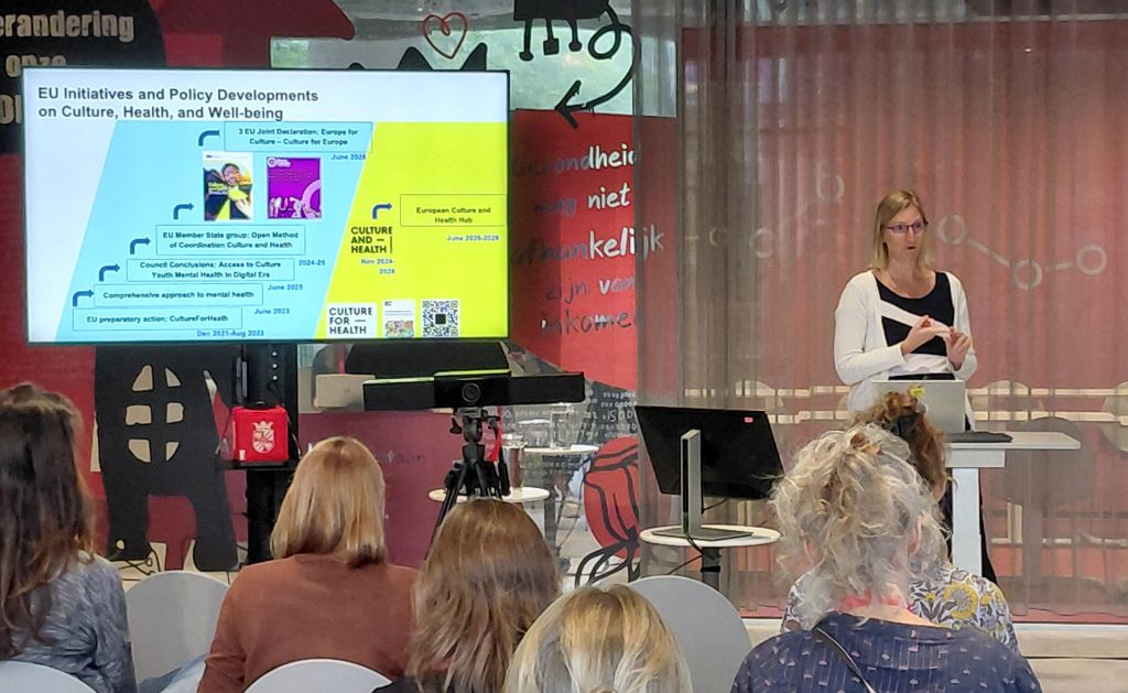 Person presenting in front of an audience. A screen with text and pictures.
