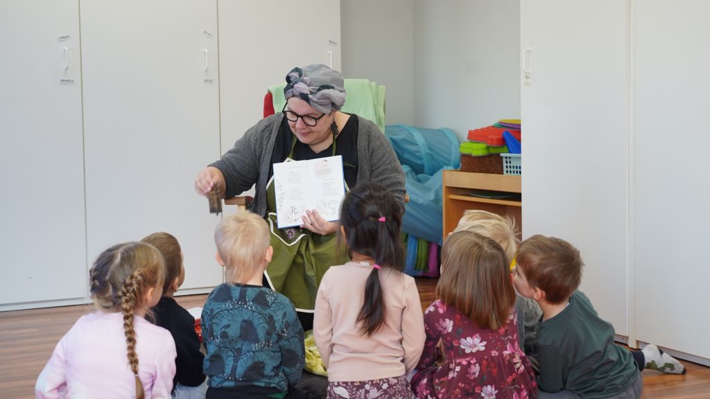 Piia Kleimola pitää toisessa kädessään lastenlorukirjaa ja toisella kädellään soittaa kelloa edessään istuville lapsille.