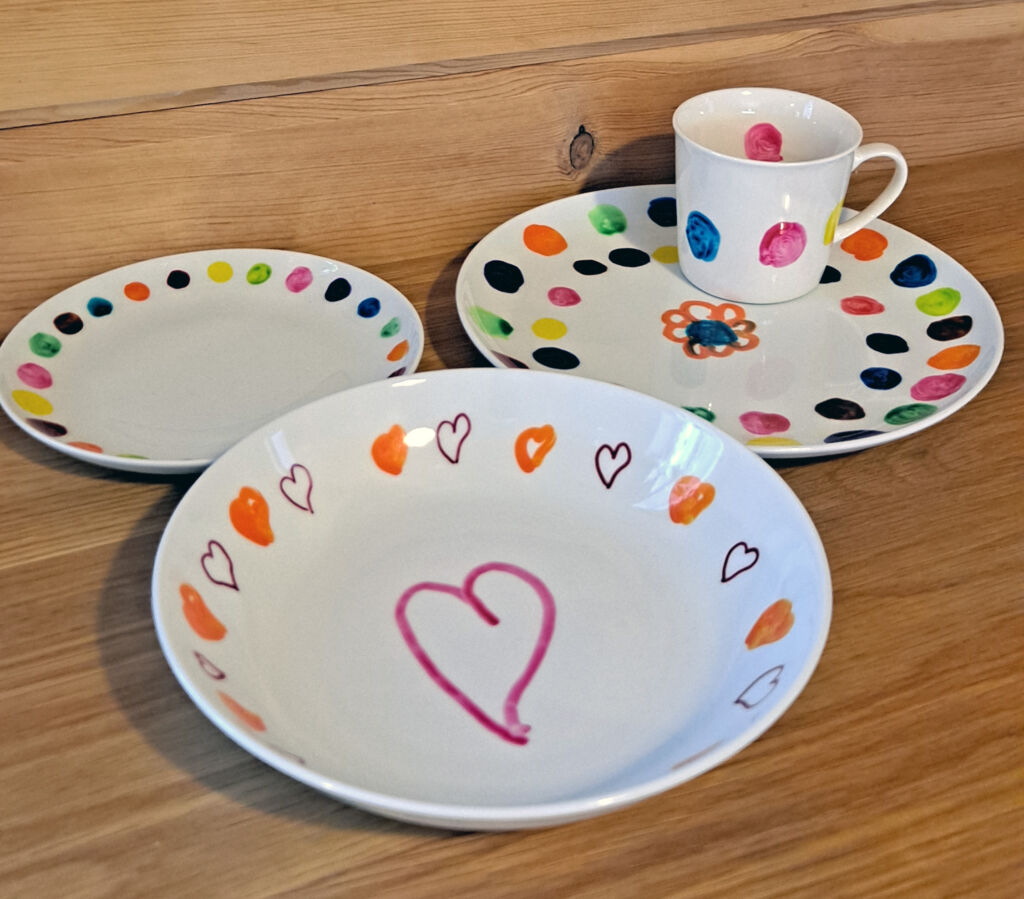 Kolme valkoista lautasta ja kahvikuppi, joihin on maalattu värikkäitä sydämiä ja palloja.