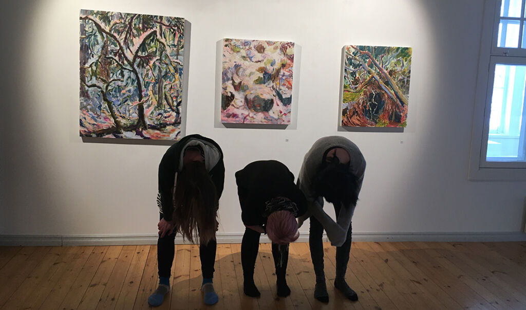 Kolme nuorta kumartaa kohti lattiaa. Taustalla on seinällä kolme taideteosta.