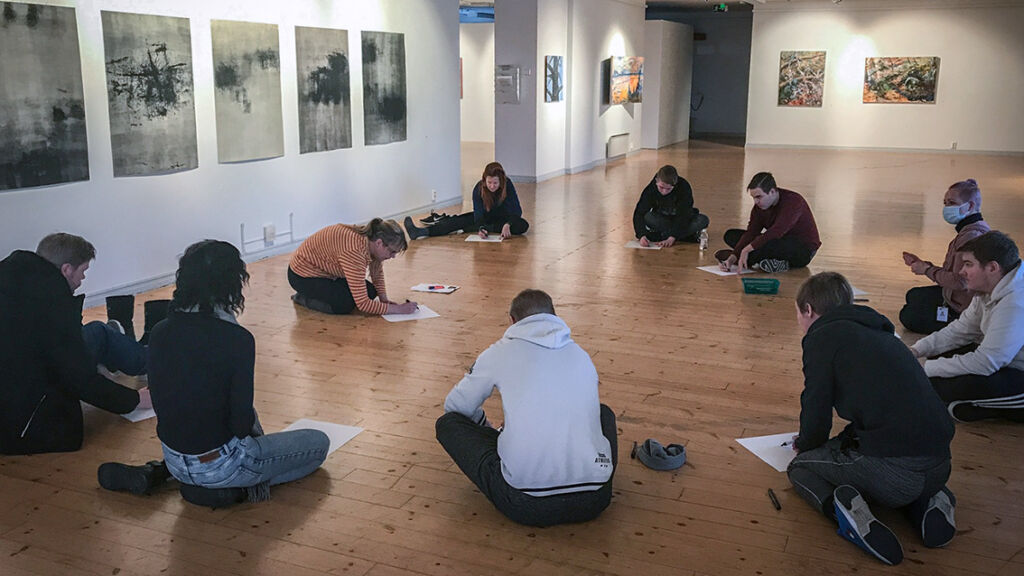 Nuoria ihmisiä taidenäyttelyssä istumassa lattialla piirtämässä.
