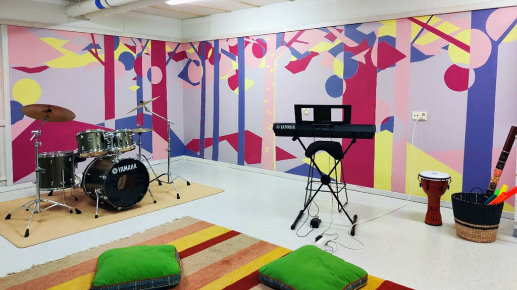 Huone, jossa on rummut, kosketinsoittimet sekä muutamia muita soittimia ja lattialla kaksi tyynyä ja matto. Seinällä on abstrahoitu seinämaalaus, joka esittää maisemaa. Maalauksen värit ovat keltainen, violetti, punainen, vaaleanpunainen ja vaaleanvioletti. 