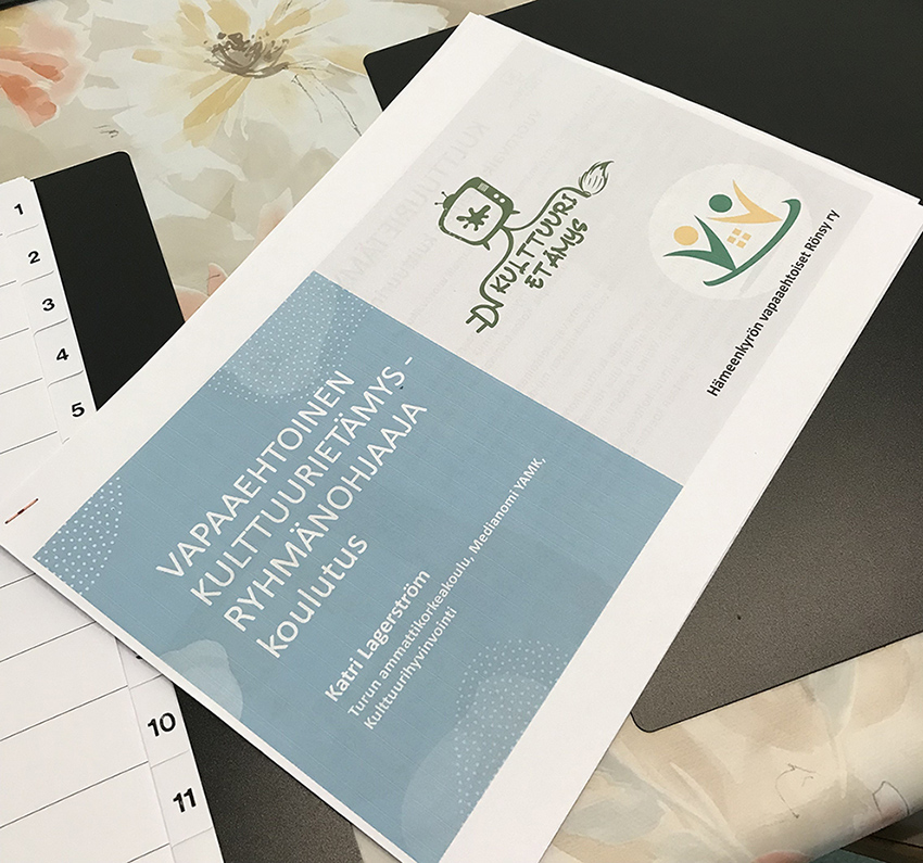 Pöydällä oleva paperinippu, jossa päällimmäisessä paperissa on teksti Vapaaehotinen Kulttuurietämys-ryhmänohjaaja -koulutus. Hämeenkyrön vapaaehtoiset Rönsy ry.