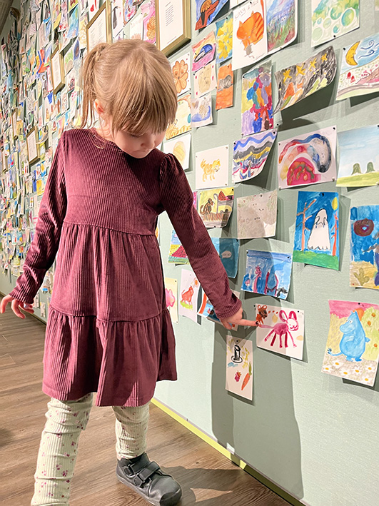 Lapsi seisoo taideteosseinän edessä osoittamassa maalaamaansa kuvaa.