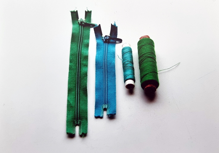 Kuvassa on kaksi vetoketjua ja kaksi lankarullaa aseteltuna vierekkäin. Toinen vetoketjuista on sininen ja toinen vihreä. Lankarullissa toistuvat samat värit.
